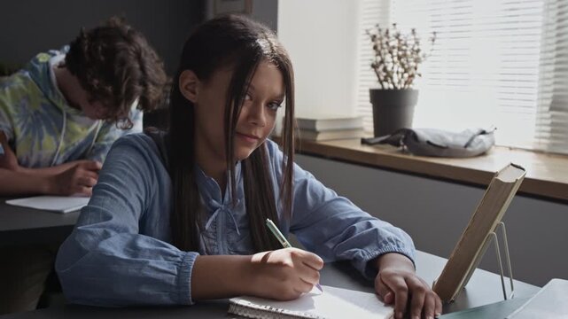 Portrait of teenage girl writing in notebook during class and looking at camera. Footage for high school promos, e-learning advertisements, and educational campaigns