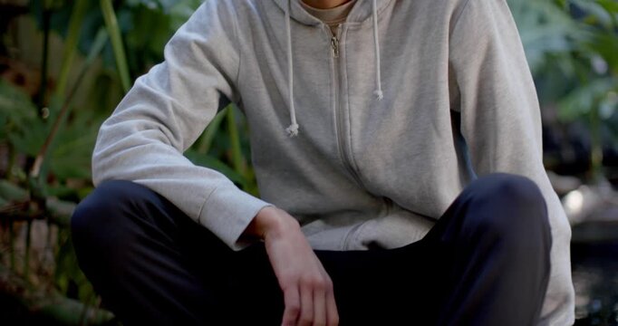Adult man wearing gray hoodie, shifting on low stone step, lifting head, smiling as camera rises