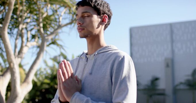 Man holding prayer pose, praying in yard while camera pulling back showing hoodie, earbud