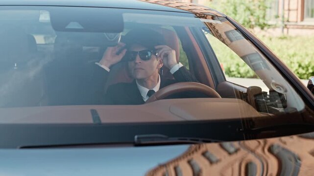Middle aged Caucasian chauffeur wearing suit and sunglasses adjusting glasses and holding steering wheel inside luxury car. Clip is perfect for promoting premium executive taxi services