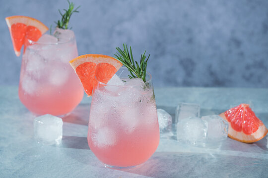 Two glasses of pink drinks sit on a table. Ice cubes float in the drinks. Slices of grapefruit and sprigs of rosemary decorate the glasses. Wellness and refreshing drink
