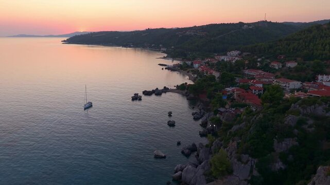 Aerial view of a yacht sailing in Aegean sea water surrounding pine forest mountains at golden hour, Loutra beach and Agia Paraskevi Pine Forest at sunset, Kassandra Peninsula