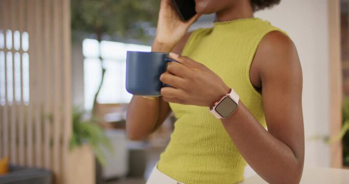 Adult African woman raising phone while answering incoming call, holding mug and laughing in office