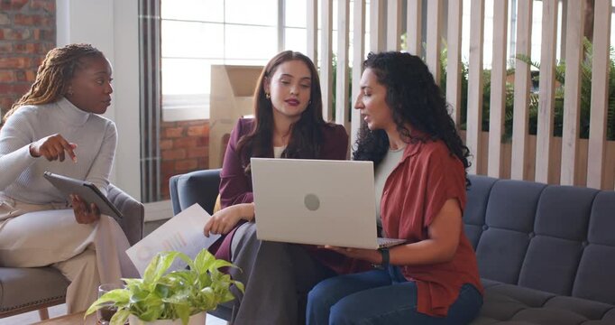 Female coworkers reviewing docs in office, left woman showing tablet, right woman scrolling laptop