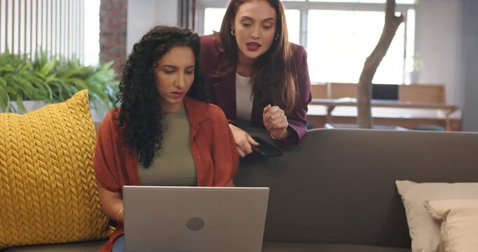 Diverse female coworkers approaching couch at office, pointing phone while guiding work on laptop