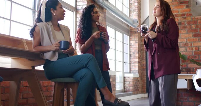 Diverse team taking break near windows, holding mugs while right woman showing camera and sipping
