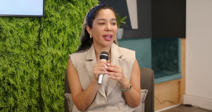Adult female speaker holding mic and gesturing at modern lounge while slide causing brief pause