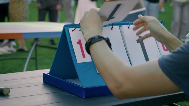 Official turns scoreboard page as players reach level score during friendly outdoor ping pong match. Close up highlights match scoring and competitive atmosphere.