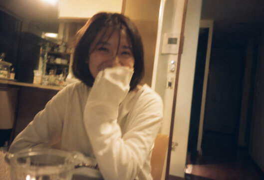 夜のダイニングで笑顔を見せる日本人女性、温かい照明とフィルムカメラ風の質感で切り取った何気ない日常ライフスタイル写真