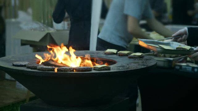 Chef lifts grilled burger patties with melted cheese from open fire grill using tongs. Street food kitchen prepares fresh burgers for hungry customers.