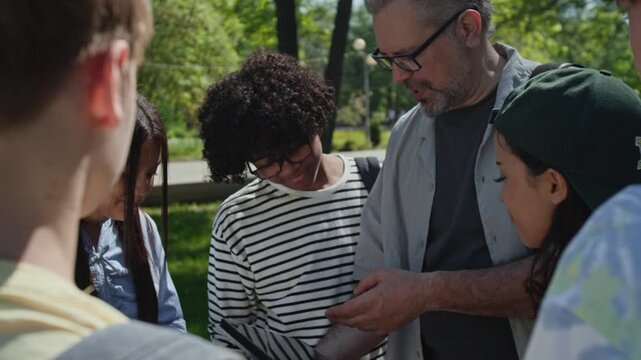 Over the shoulder view of middle aged teacher explaining lesson on tablet to diverse group of teenagers outdoors in park. Clip for promoting outdoor education and interactive learning programs