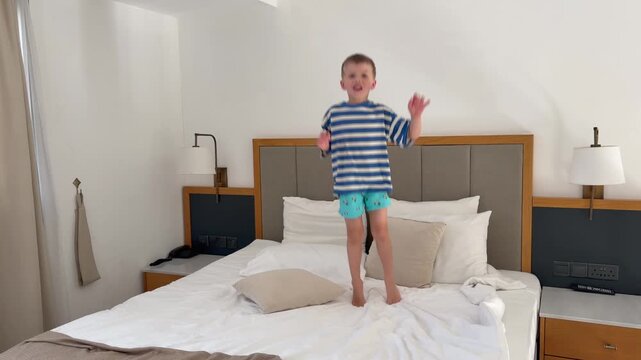 Little boy jumping on hotel bed with happy smile and playful energy. Family vacation, carefree childhood, travel lifestyle, excitement and memorable holiday moments concept.