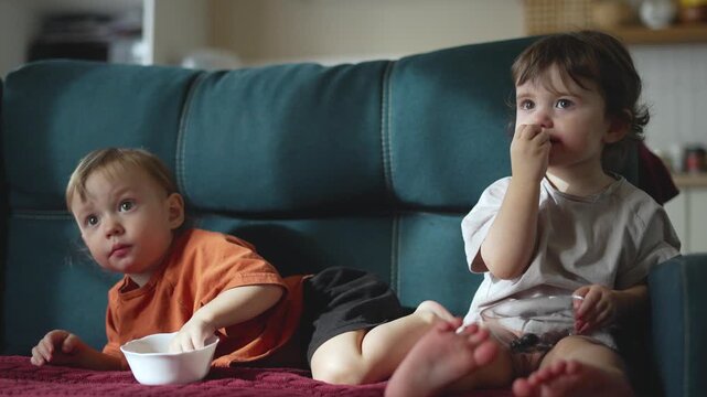 Two children sitting on couch making facial expressions while snacking. Kids react emotionally watching together. Boy and girl show reactions. Children express emotions. Kids snack and react.