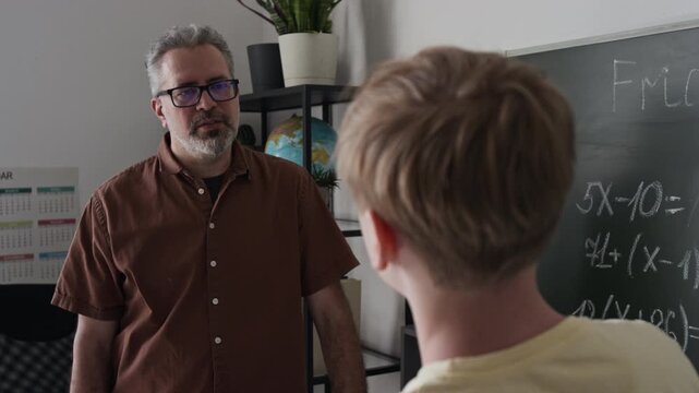Middle aged teacher explaining math equation to teenager writing on blackboard. Clip for e-learning platforms and high school promo videos