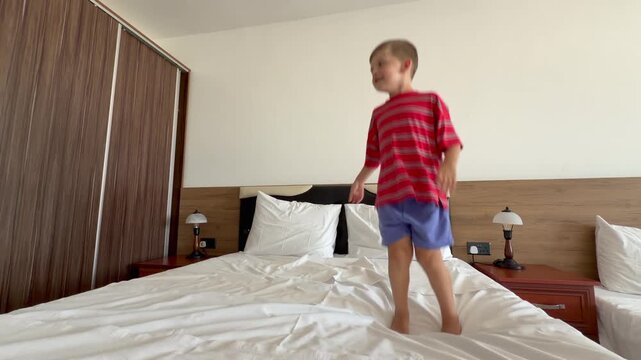 Little boy happily bouncing on soft hotel bed during family vacation. Bright modern interior, joyful childhood, active movement and relaxing holiday atmosphere concept. Child smiling jumping on bed
