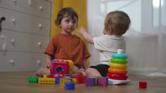 Two toddler children playing together on the floor with colorful educational toys. Boys interact while sharing building blocks and rainbow stacking rings in a bright modern playroom setting.