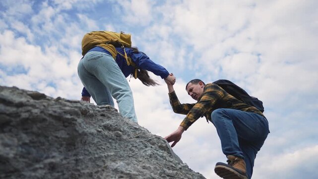 Hiker helps partner climb rocky mountain terrain outdoors. Man assists woman climbing rock during hiking adventure. Couple shows teamwork support during outdoor climb. Hikers help each other.