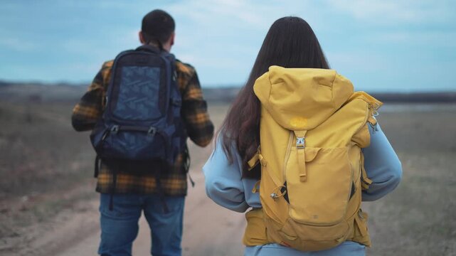 Young couple backpackers walking on empty desert road during hiking adventure. Man waving while woman with yellow backpack follows. Travelers explore wilderness trail on outdoor expedition journey.