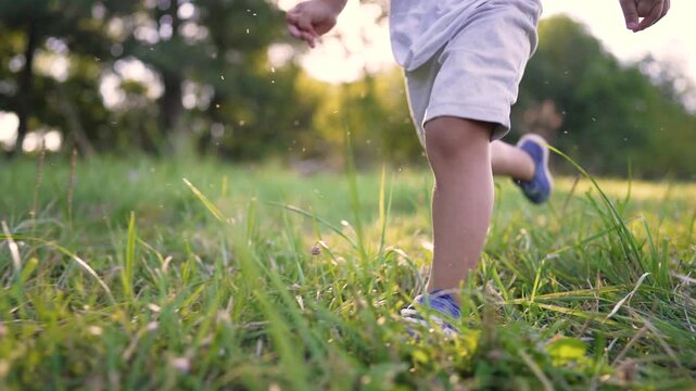 Child running through grass field at sunset. Boy plays in park wearing sneakers. Kid runs on green meadow outdoors. Active child enjoys nature. Boy sprints across grass in evening sunlight.