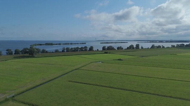 Aerial view flying forward and upward over green fields with drainage ditches, with Markermeer and distant wind turbines on the horizon.