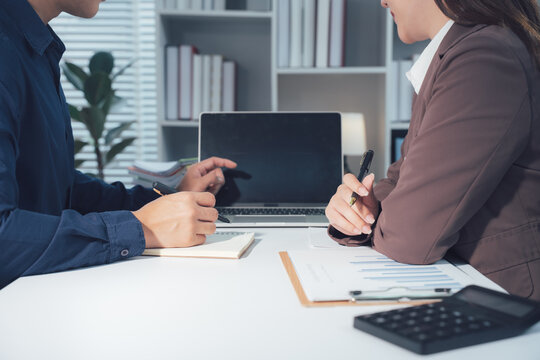 Business Meeting: Two professionals in a modern office setting engage in a focused discussion over a laptop, papers, and a calculator, signifying collaboration and strategic planning.