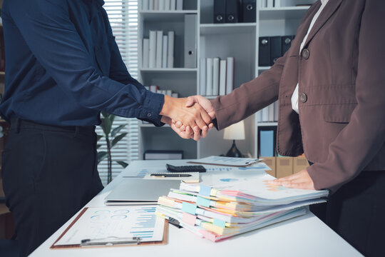Successful Business Partnership Handshake: Two business people seal a deal with a firm handshake, signifying trust and collaboration in a modern office setting with stacks of documents on the desk.