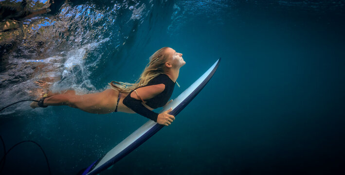 Dynamic underwater view of blonde woman paddling surfboard to catch wave