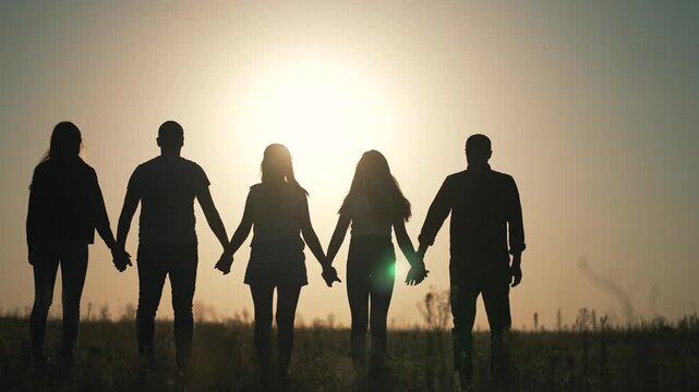 Group of friends holding hands in silhouette against sunset sky outdoors. Five people stand together showing unity and friendship. Friends create human chain at golden hour. Teamwork concept.