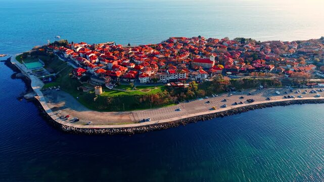 Drone view of Nessebar peninsula coastline, Bulgaria. Elevated shot of red roofed old town, tennis court and coastal road along the rocky breakwater shoreline.