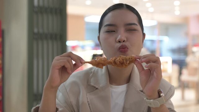 Hungry young Thai Isan businesswoman eating a fried chicken skewer at a food court table