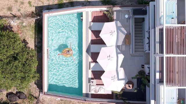 Young woman enjoying sunbathing sitting on inflatable tube in private pool at boutique hotel and resort, Rooftop terrace swimming pool and sun loungers, Old Square of Pefkochori, Overhead aerial view