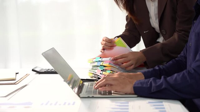 Examining Financial Documents: Two colleagues meticulously review a stack of colorful financial reports and documents while working together on a laptop.