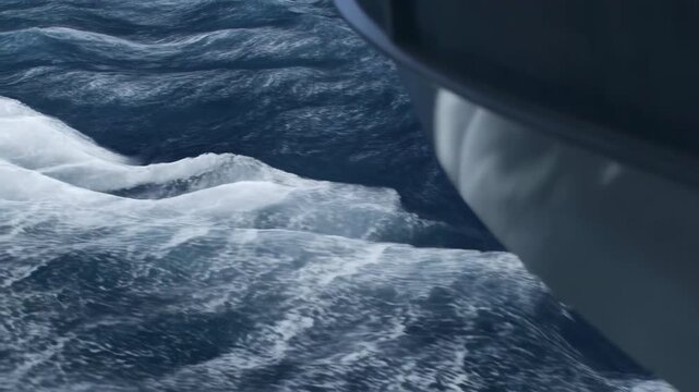 Dark blue ocean beside vessel, white foam streaming across rough waves from low side view, showing blurred hull edge, cold maritime travel mood and textured open sea surface