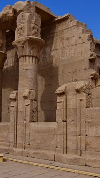 Ornate colonnade with carved floral capitals and hieroglyphic screen walls at Edfu Temple, Egypt. Ptolemaic architecture against vivid blue sky. Travel and history content