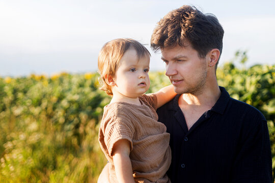 Happy father walking with son in sunny summer field. Stylish dad holding baby boy, relaxing on background of sunflowers. Family leisure with a child