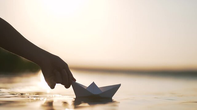 Hand releases paper boat floating on calm water at sunset. Origami vessel drifts across peaceful lake surface. Golden hour light reflects warmly. Paper ship sails in evening glow softly.