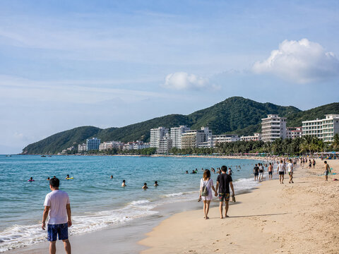 Visitors wading along a broad Hainan beach with resort hills in the distance.generative AI