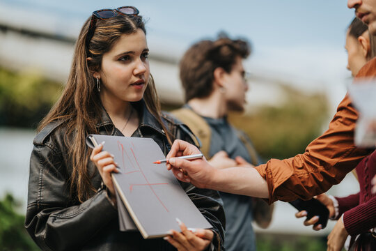 High school students stand outdoors while one student holds a clipboard and another writes with a pen. A small group discusses and collaborates on a survey or project in a casual setting.
