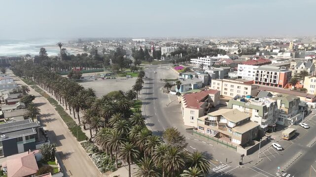 Cinematic aerial footage of the coastal desert city of Swakopmund, Namibia, featuring palm trees and a road curving along the Atlantic beachfront.