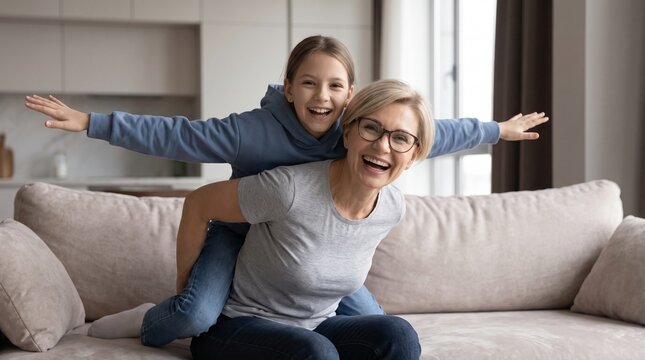 Playful senior blonde grandmother in glasses giving a piggyback ride to her happy little granddaughter, pretending to fly with arms outstretched like an airplane on a sofa at home.