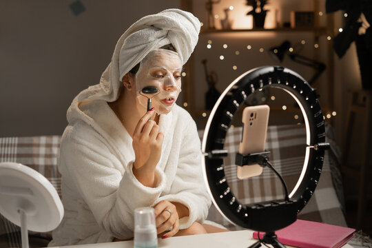 Young female blogger wearing white bathrobe and towel live broadcasting cosmetic and skincare tutorial content in front of ring light in the evening at home. Beauty, blogging and skincare