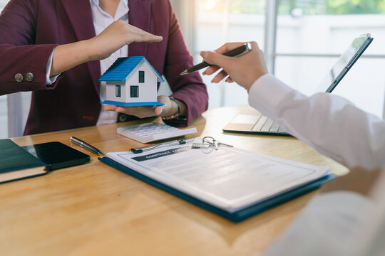 A real estate agent holding a small house model while a client signs a contract, and a home insurance agent using their hands to protect the house model.