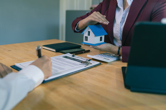 A real estate agent holding a small house model while a client signs a contract, and a home insurance agent using their hands to protect the house model.