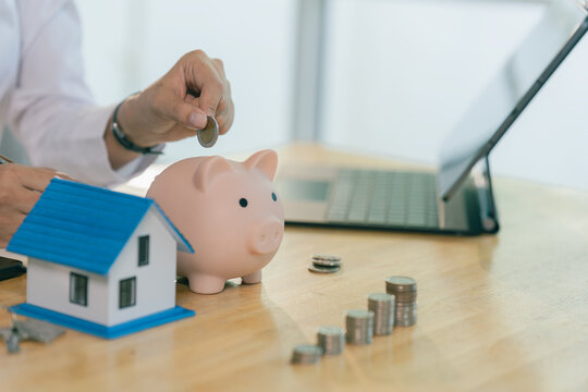 A smiling Asian woman is dropping a coin into a piggy bank to save up for a home, saving money for future real estate investment.
