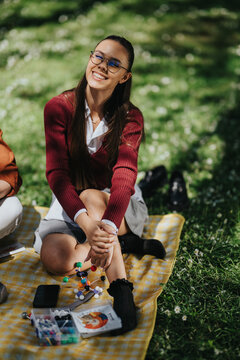High school student in uniform smiles while sitting on a picnic blanket with study materials. A cheerful teen enjoys outdoor studying with a molecular model and supplies on grass.