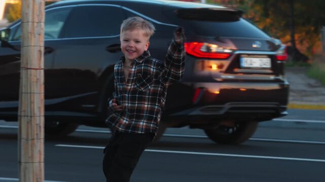 Young boy in plaid shirt joyfully runs alongside a road, with a black SUV and construction site visible in the background