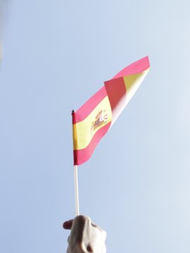 A close-up of a hand waving a miniature Spanish flag with a bright blue sky in the background.