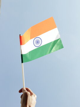 A close-up of a hand waving a miniature India flag with a bright blue sky in the background.