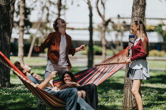 A group of young friends relaxing and laughing in a striped hammock between trees. The scene shows playful conversation and carefree leisure in an urban park setting.