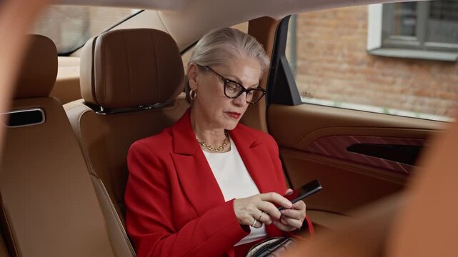 Senior Caucasian businesswoman sitting in back seat of luxury car, taking smartphone out of purse and answering call. Video suitable for corporate communication and executive travel marketing